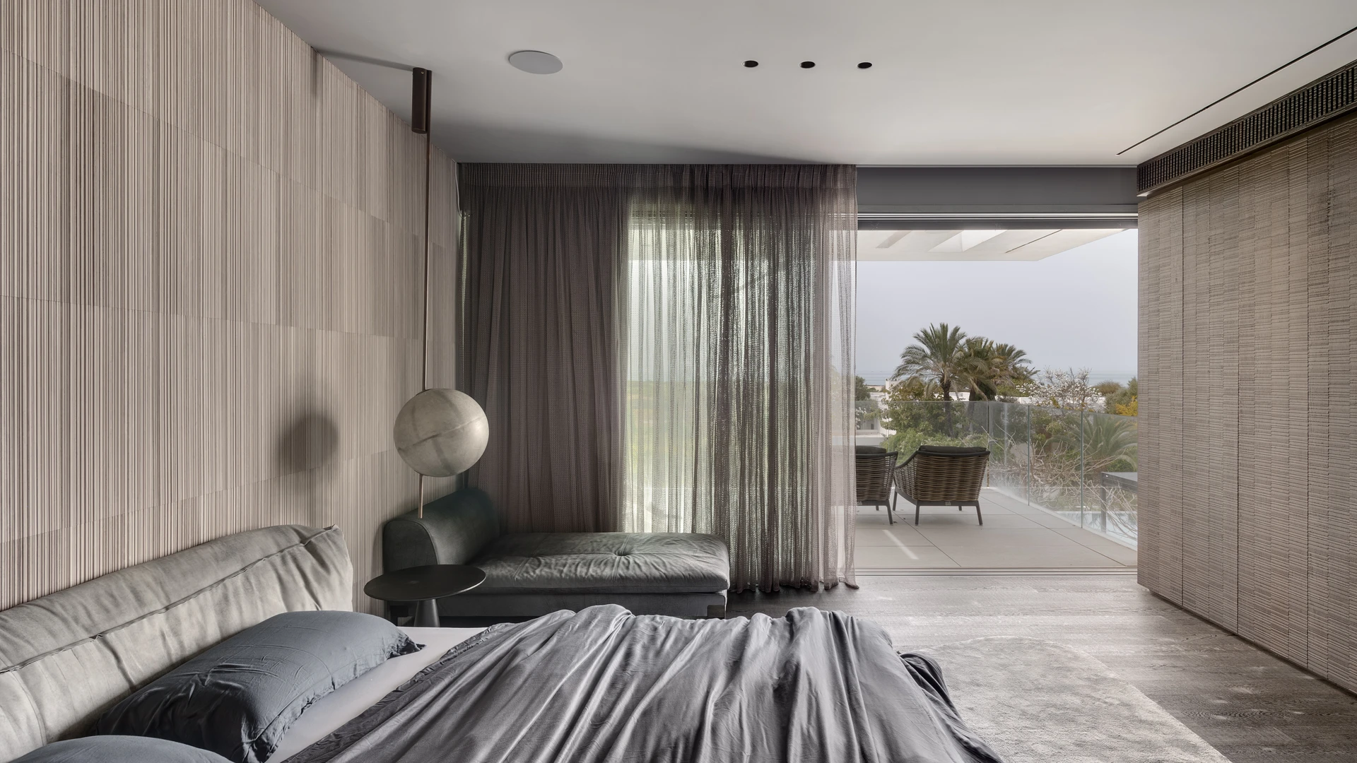 Contemporary bedroom with textured wallpaper and balcony access overlooking trees and outdoor lounge chairs.